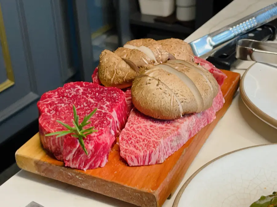Spessa carne di manzo cruda e funghi shiitake affettati sottilmente disposti su un tagliere di legno con del rosmarino