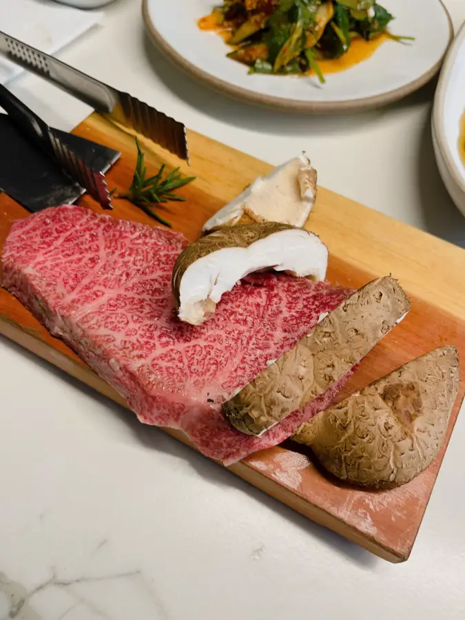 Carne di manzo cruda con evidente marezzatura e funghi shiitake affettati disposti su un tagliere di legno