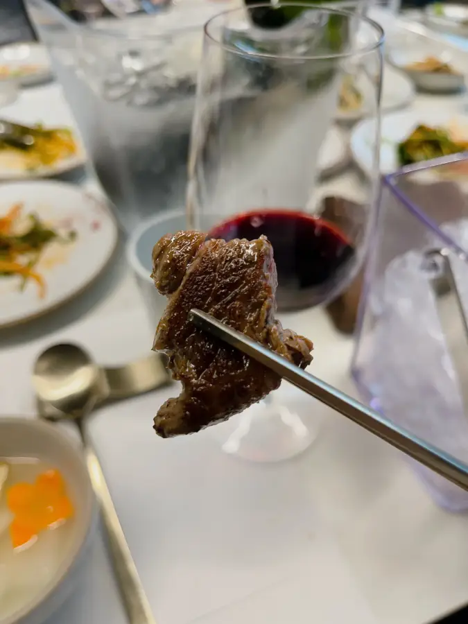 Un pezzo di carne di manzo ben grigliata sollevato con le bacchette, con un bicchiere di vino visibile sullo sfondo