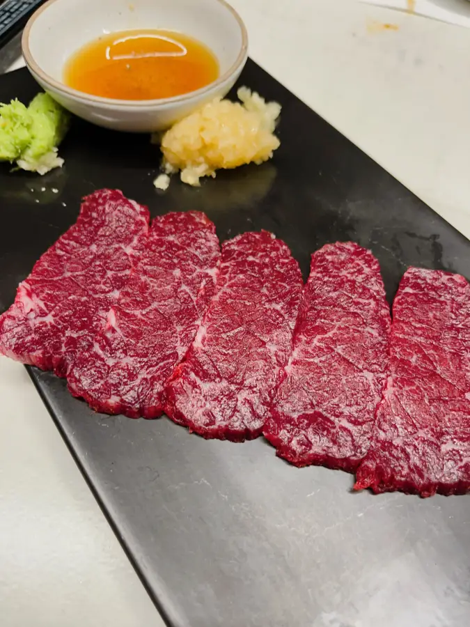 Cinque fette di carne cruda rossa disposte ordinatamente su un piatto nero con salsa e wasabi
